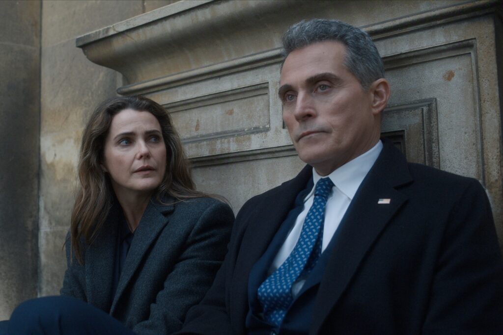 Nuværende billede: "Two people in formal coats sit on outdoor stone steps, appearing serious and deep in conversation, with an old stone wall in the background. The man wears a suit and tie with an American flag pin, suggesting a political or official context."