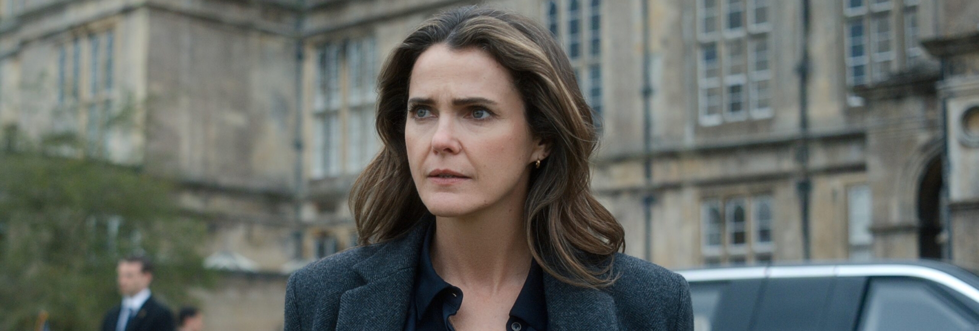 "A woman with wavy brown hair in a dark coat stands outside a grand historic building, looking serious. There is a black car and security personnel in the background, suggesting a formal or official environment."